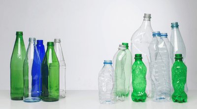 HDPE vs PET bottles comparison showing clear, green, and blue plastic bottles beside glass bottles, illustrating material, texture, and recyclability differences in sustainable packaging.